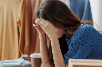 Eine Frau stützt den Kopf vor dem Laptop erschöpft in ihre Hände, offenbar mit Kopfschmerzen. Im Hintergrund ist eine Kleiderstanke mit mehreren Oberteilen zu sehen.