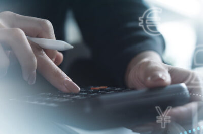 Ein professioneller Arbeitsbereich mit Fokus auf Finanzanalyse. Im Vordergrund tippt eine Person mit einem Stift in der Hand auf einem Taschenrechner, wobei ihre Hände scharf im Fokus sind. Im Hintergrund verschwimmen visuelle Darstellungen von Währungssymbolen und Aufwärtstrends auf Diagrammen, die die globale Finanzwirtschaft symbolisieren.