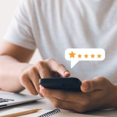 „Eine Nahaufnahme einer Person, die ein Smartphone in der Hand hält, während sie darauf tippt. Neben dem Smartphone schwebt eine Sprechblase mit fünf Sternen, die eine Bewertung darstellt. Im Hintergrund ist ein Laptop und ein Notizbuch zu sehen, die auf einem Schreibtisch liegen.“