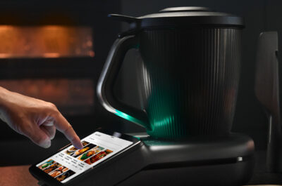 Der neue Thermomix TM7 mit großem Touchscreen: Eine Person tippt auf das Display eines schwarz-matten Thermomix TM7, während der beleuchtete Mixtopf auf der Basis steht. Im Hintergrund ein Backofen und frische Zutaten auf der Arbeitsplatte.