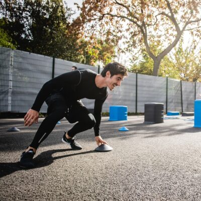 Ein junger Mann in schwarzer Sportkleidung bewegt sich mit einem Lächeln durch einen Outdoor-Fitness-Parcours. Er greift nach einem kleinen Kegel, während andere Teilnehmer im Hintergrund zwischen blauen und grauen Hindernissen trainieren. Die Szene spielt sich auf einem sonnigen, von Bäumen umgebenen Sportplatz ab.